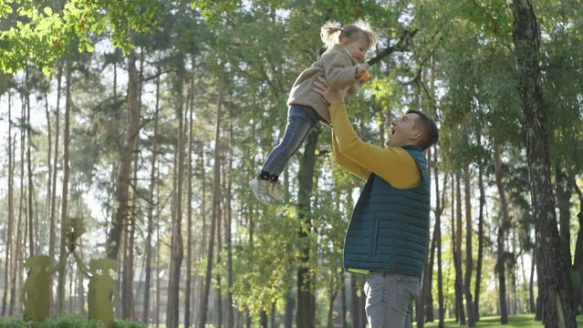Father throws his little daughter up, leaves fall from trees in the park, autumn. Sunny weather in nature, a man throws a girl over his head, a happy family. High quality 4k footage