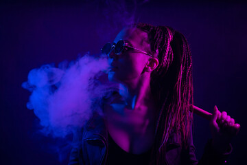A young woman with dreadlocks smokes a cigarette in a club. Wearing black glasses, he holds a painless bat in his hands. Girl with afro pigtails in the smoke at night in neon color. Scoundrel