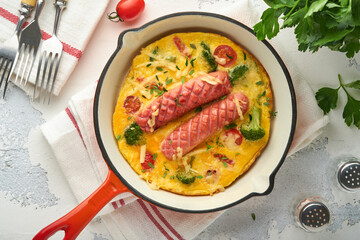 Omelette or frittata with two sausages, melted cheese, cherry tomatoes, broccoli and parsley on red skillet or iron pan on white old concrete background. Healthy breakfast food concept. Top view.
