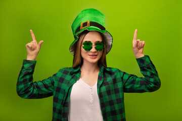 Happy smiling young woman wearing checkered plaid shirt, green leprechaun hat with clover shaped glasses and pointing index fingers up with two hands on green background.

St Patricks Day.