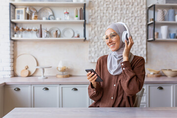 Portrait of a young Muslim woman in a hijab sitting at home in the kitchen wearing headphones and...