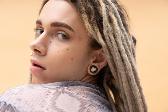 Portrait of young nonbinary person with tattoo and dreadlocks posing isolated on yellow.