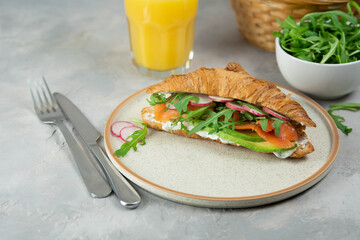 Croissant sandwich with cream cheese, salmon and arugula on a white plate, gray background, close-up. Healthy breakfast concept.