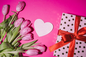 Love card in shape of heart among spring pink tulips flowers and pink gift box with red ribbon on pink background.