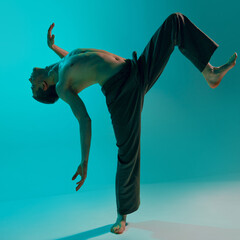 Contemporary, modern dance style. Young flexible shirtless man dancing experimental dance over blue, cyan studio background. Concept of art, body aesthetics, motion, action, inspiration.