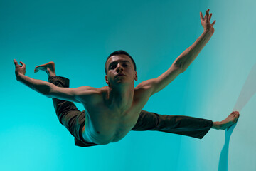Contemporary dance style. Young shirtless man dancing over blue, cyan studio background. Expressive...