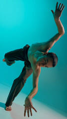 Contemporary dance style. Young shirtless man dancing over blue, cyan studio background. Flexile...