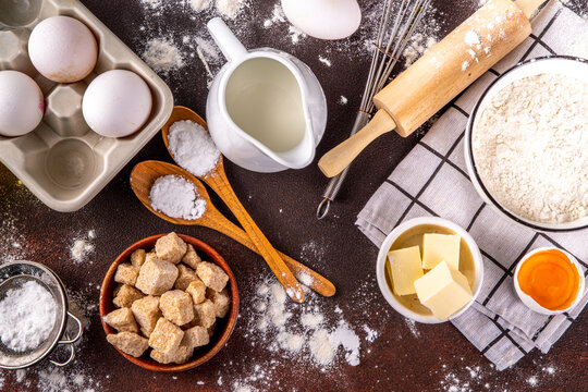 Baking ingredients background. Cooking ingredients (flour, eggs, milk, brown sugar, butter) with utensils on dark brown background top view copy space - Powered by Adobe