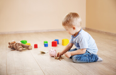 Naklejka premium Cute boy sitting on the floor indoor and inserting euro coins in piggybank.