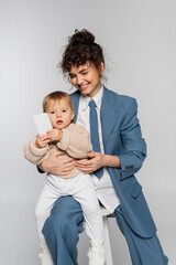 cheerful businesswoman in blue suit sitting on chair while toddler daughter holding smartphone isolated on grey.