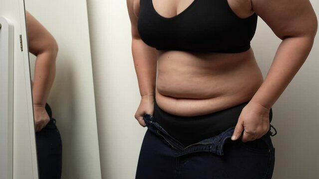Asian oversize woman pulling jeans trousers up, in front of a mirror. Excess belly fat, Unhealthy lifestyle, Diet concept.