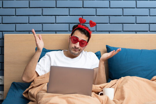 Upset man lying in the bed and talking on video call on his laptop with his sweetheart at the Valentine's Day. He wearing funny accessories but feeling bad because of loneliness - Powered by Adobe