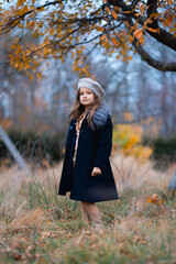 A lovely little girl with a hat and blue coat is standing in an autumn garden and looking at the camera