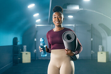 Young African American woman in sportswear standing in gym, carrying rolled yoga mat and bottle of water.