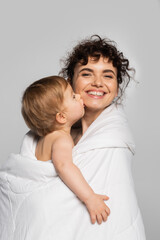 toddler baby girl kissing cheek of positive mother covered in white duvet isolated on grey.