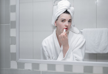 young brunette paints her lips with red lipstick, looking in the mirror