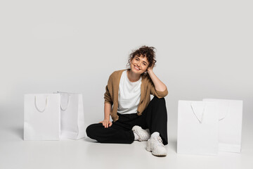 full length of cheerful woman in casual clothes sitting near shopping bags on grey.