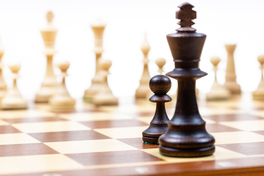 black pawn and queen against white chess pieces in background on wooden chessboard close up (focus on the black pawn)