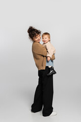 full length of happy mother in casual clothes holding in arms cheerful baby daughter in sweater on grey.