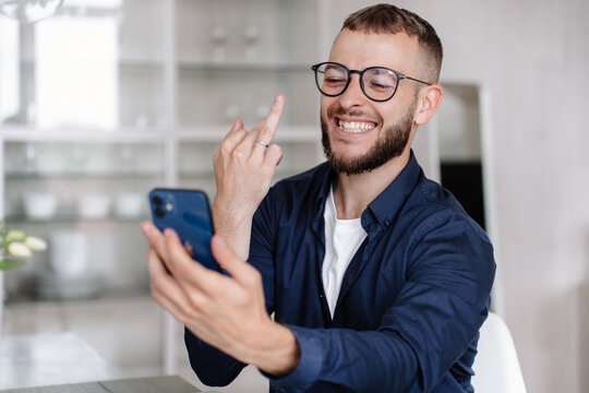 Cheerful caucasian young man sitting at desk makes video call by phone shows to friend ring finger with wedding ring on it. Happy newlywed guy shares happiness with parent at conversation by cellphone - Powered by Adobe