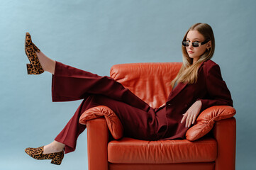 Fashionable confident woman wearing elegant marsala color suit, sunglasses, leopard print loafer shoes, posing, sitting in brown leather armchair. Studio fashion portrait. Copy. empty space for text - 565372808
