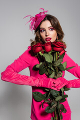 brunette young woman in magenta color gloves and hat with feather holding red roses on grey.