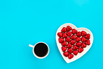 Heart shaped plate with heart shaped red foil chocolates and coffee cup on a blue background, flat...