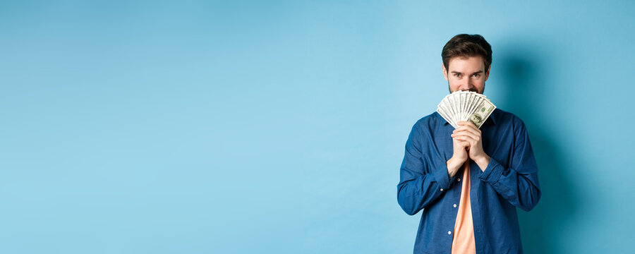 Young Man Hiding Smiling Face Behind Money And Looking Determined At Camera, Standing On Blue Background