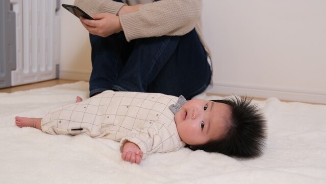 育児につかれた親がスマートフォンを眺めて育児から逃避している様子の写真。乳児の赤ちゃんは近くで寝転がったまま放置されている