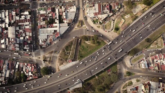A dynamic top-down ascending aerial shot above a busy highway on top of a roundabout. Lots of vehicles are passing by the Plaza de los Virreyes subway train station.