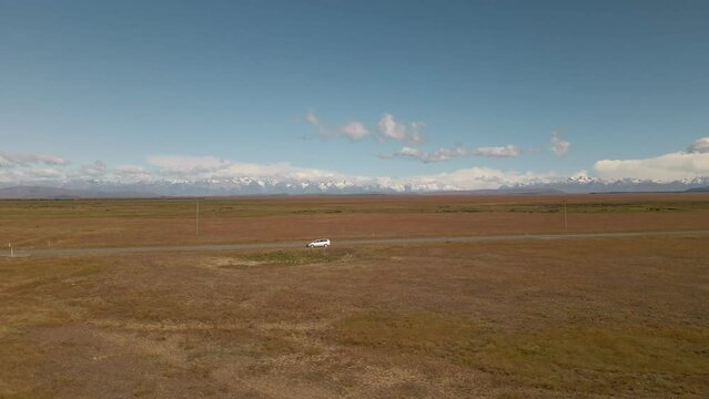 Silver car on side of picturesque road within dry plains with snow covered mountains in backdrop