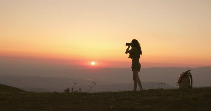 Inspired and passionate about work of photographer, woman stands on very edge of hill and takes photo of red misty dawn in early morning. Dark silhouette of woman photographer in warm summer sunrise. 