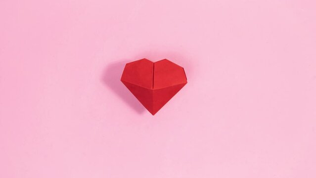 4k Small red heart appears between fingers of woman's hand, which transforms into origami heart. Pink background. Concept of valentine's, mother's day holiday, wedding.  Stop motion animation.