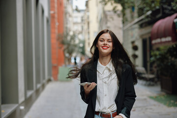 Business brunette woman with red lips smile with teeth with a phone in her hands, white shirt and black jacket fashion on the street, summer trip, vacation in the city tourist