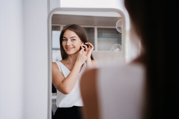 Fototapeta premium Attractive young Italian woman standing at mirror putting in earrings toothy smiles prepares for date. Cheerful caucasian girl at sunny morning at home. Pretty model on vacations at hotel room.