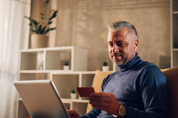 Middle aged man using a laptop and a credit card while sitting on a sofa at home