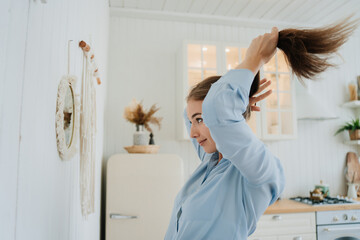 Attractive blonde Italian young woman in blue shirt stands at mirror makes ponytail against blurry...