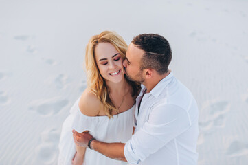 A tanned young caucasian bearded guy kisses a beautiful fun smiling blonde woman against the backdrop of the setting sun. Desert, sandy beach, rest and relaxation. Concept of honeymoon.