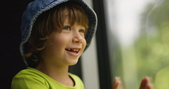 Cute little boy looks at views from bus window with admiration and delight. Child boy smiles, leaning on the glass of window with his hands and considers sights of unfamiliar city to which he arrives