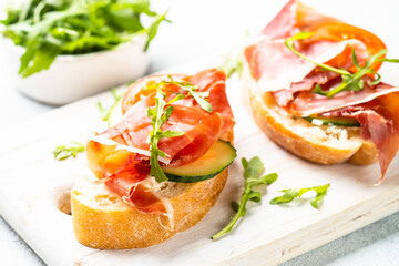 Appetizer crostini, open sandwiches with cream cheese, prosciutto, cucumber and arugula at white table.