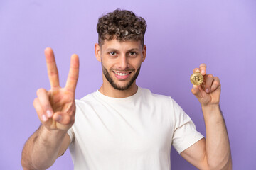 Young caucasian man holding a Bitcoin isolated on purple background smiling and showing victory sign