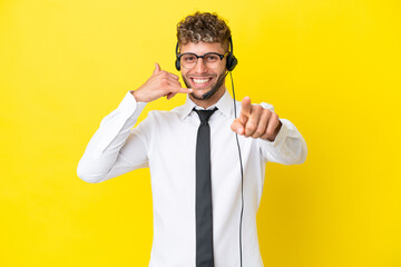 Telemarketer blonde man working with a headset isolated on yellow background making phone gesture and pointing front