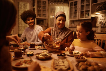 Happy Muslim mature woman serving food to her family during meal at dining table.