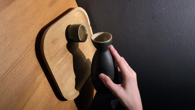 Vertical: Female hand pouring japanese sake from jug into a black ceramic cup