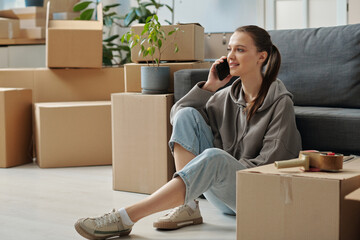 Young woman discussing her move with loaders by the mobile phone while sitting on the floor among packed boxes
