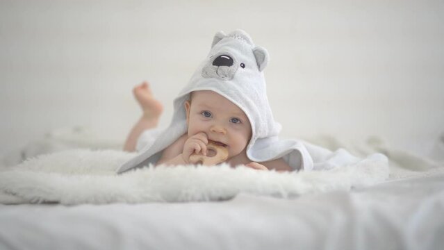 A cute funny little baby with a wood teether is resting in bed after bathing, covered with a towel, laughing happily. The concept of a baby bath. Copy space. Soft focus.