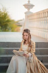 A beautiful young woman in an eighteenth century historical gold dress stands on the stairs of a mansion with a book in her hands. the princess is reading in the palace.