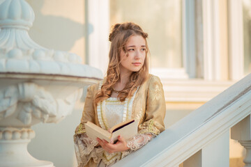 A beautiful young woman in an eighteenth century historical gold dress stands on the stairs of a...