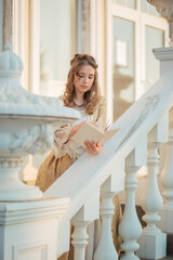 A beautiful young woman in an eighteenth century historical gold dress stands on the stairs of a mansion with a book in her hands. the princess is reading in the palace.
