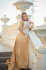 A beautiful young woman in an eighteenth century historical gold dress stands on the stairs of a...